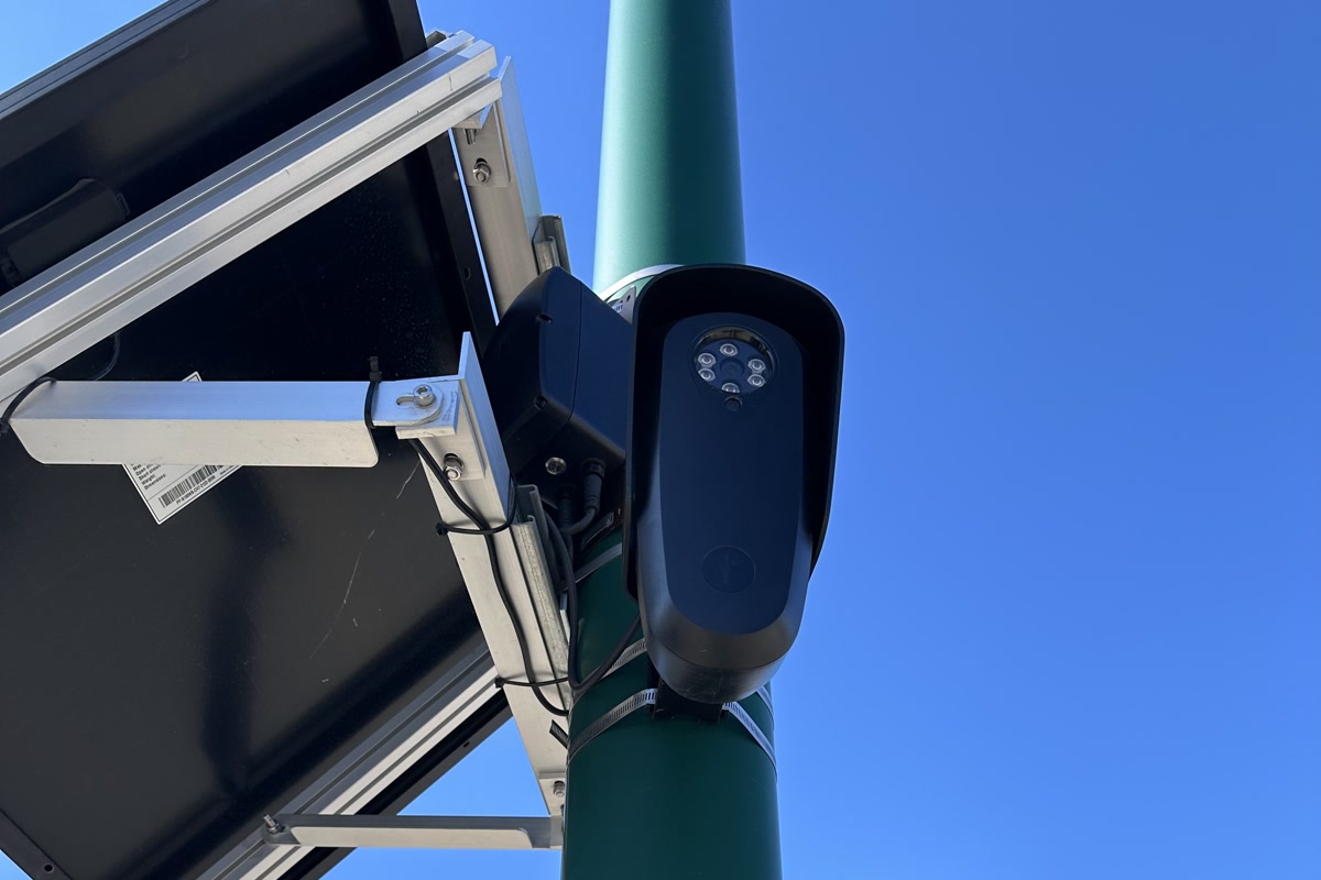 Close-up view of a Flock Safety automated license plate reader mounted above a road sign against a blue sky.